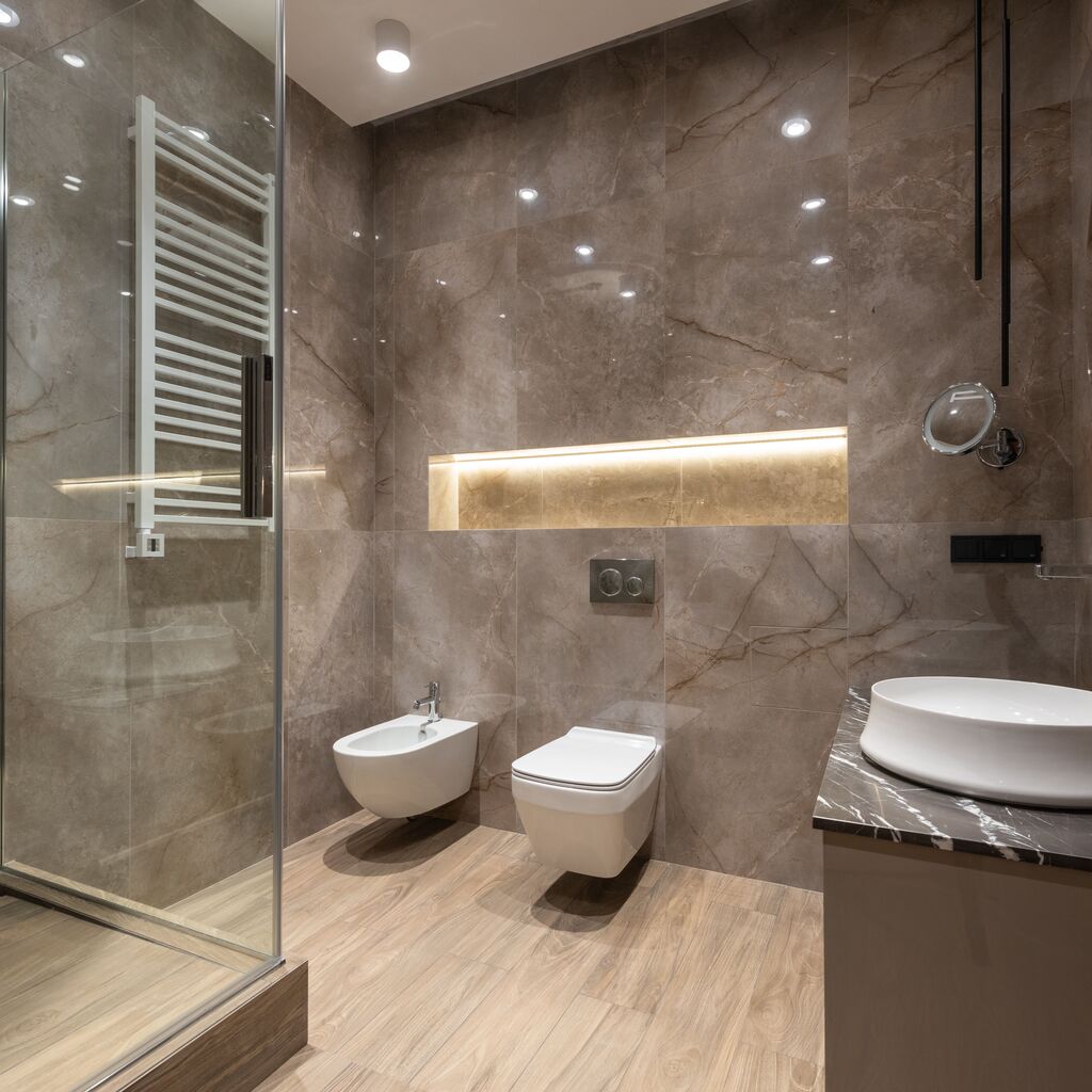 Brown tiled bathroom with wooden floors and two white toilets, a clear shower, and a white basin.